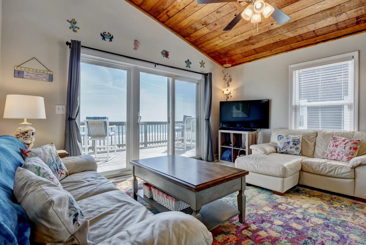 A cozy living area with comfortable seating, colorful area rug, and large windows that offer a view and plenty of sunlight. The atmosphere is perfect for relaxing after a day at the beach or planning the next adventure.