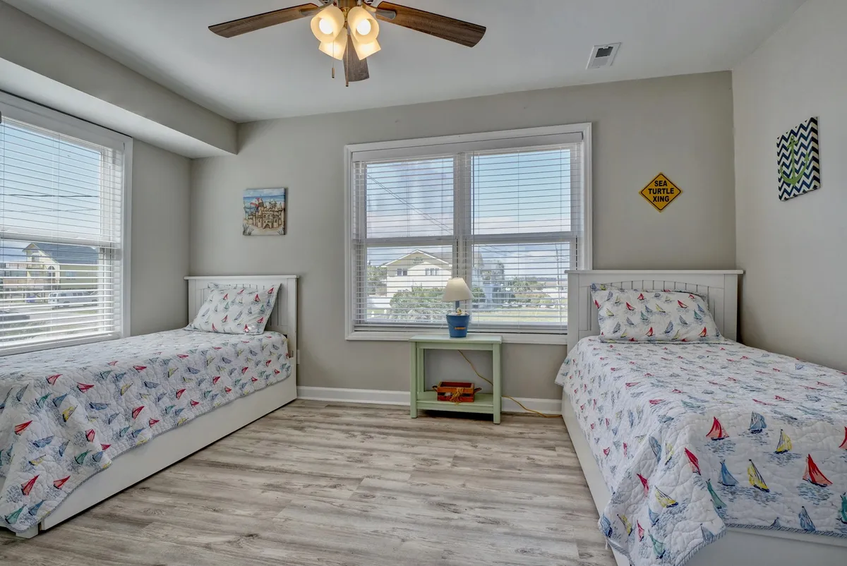 A playful twin bedroom with two nautical-themed twin beds, a small green side table with a lamp, and large windows filling the room with natural light. The space feels inviting and ideal for children or extra guests, suggesting both fun and comfort.