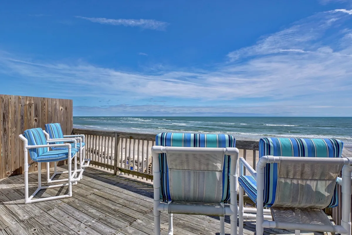 The image features a serene oceanfront deck overlooking the sweeping shoreline of North Topsail Beach. Beach chairs are arranged invitingly on the deck, framed by a white railing, with the endless blue of the Atlantic stretching out beyond. The scene suggests relaxation, with gentle waves, salty breezes, and the promise of stunning sunrises and starlit nights.