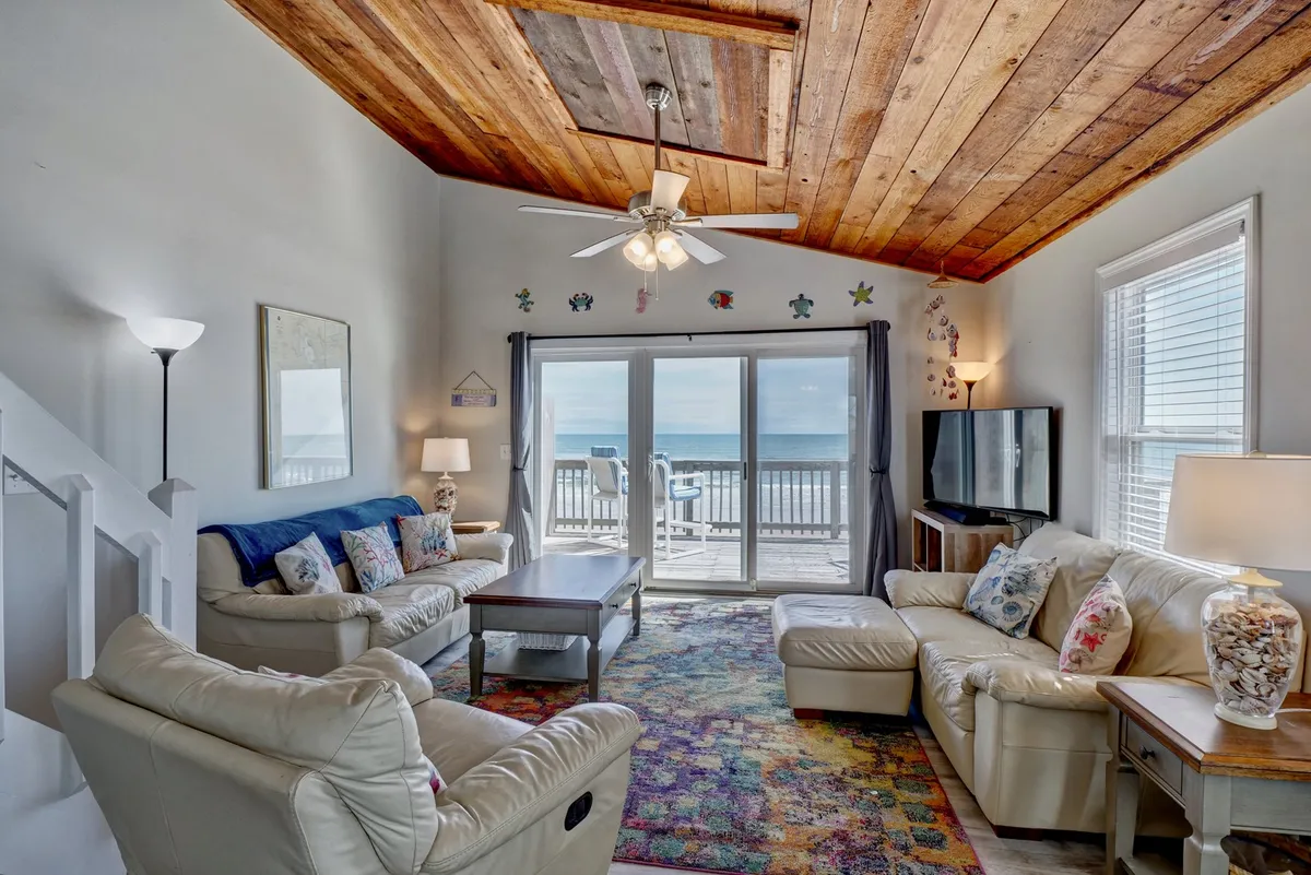 The focus is on a welcoming living area with large sliding glass doors that frame a breathtaking ocean view. A colorful area rug and wooden ceiling add warmth and character. The setup encourages relaxation, conversation, and entertaining, with the ocean always in sight.