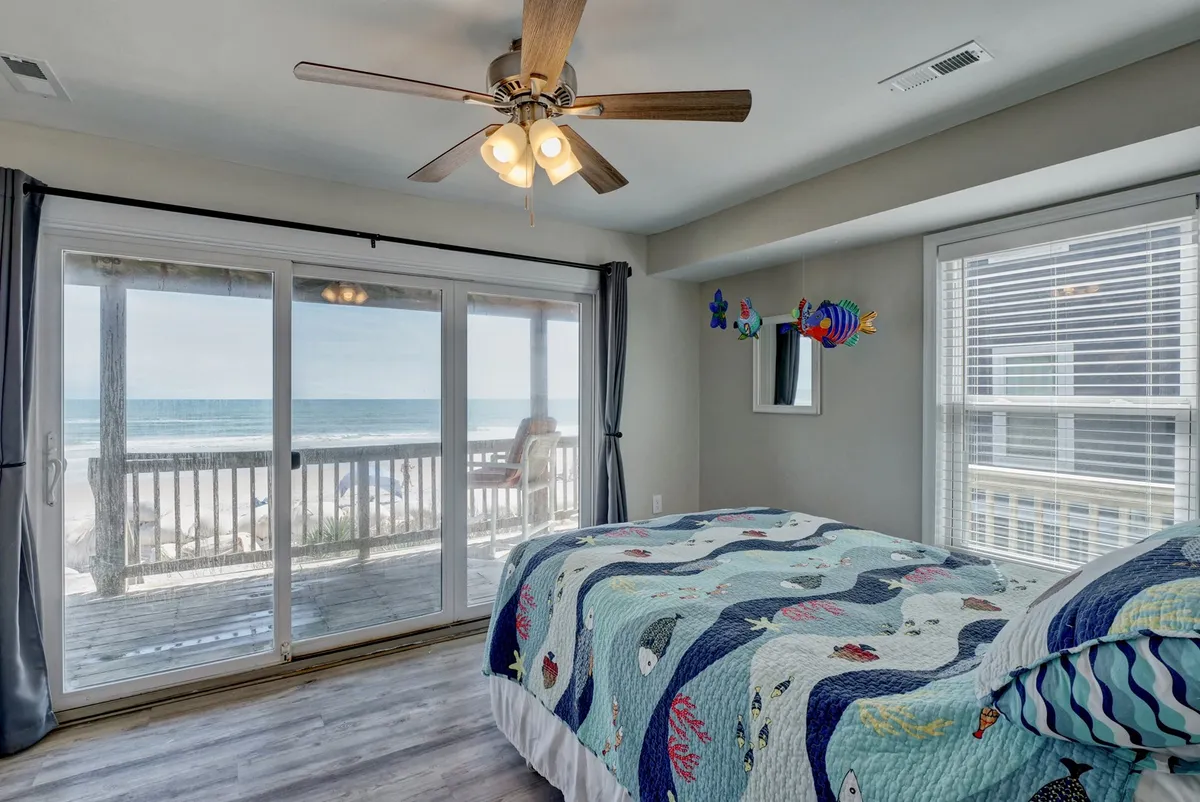 A bright queen bedroom featuring a colorful quilt on the bed, large sliding glass doors that open directly onto a private deck, and a sweeping view of the ocean beyond. The space is awash in natural light, inviting relaxation and easy access to the outdoors.