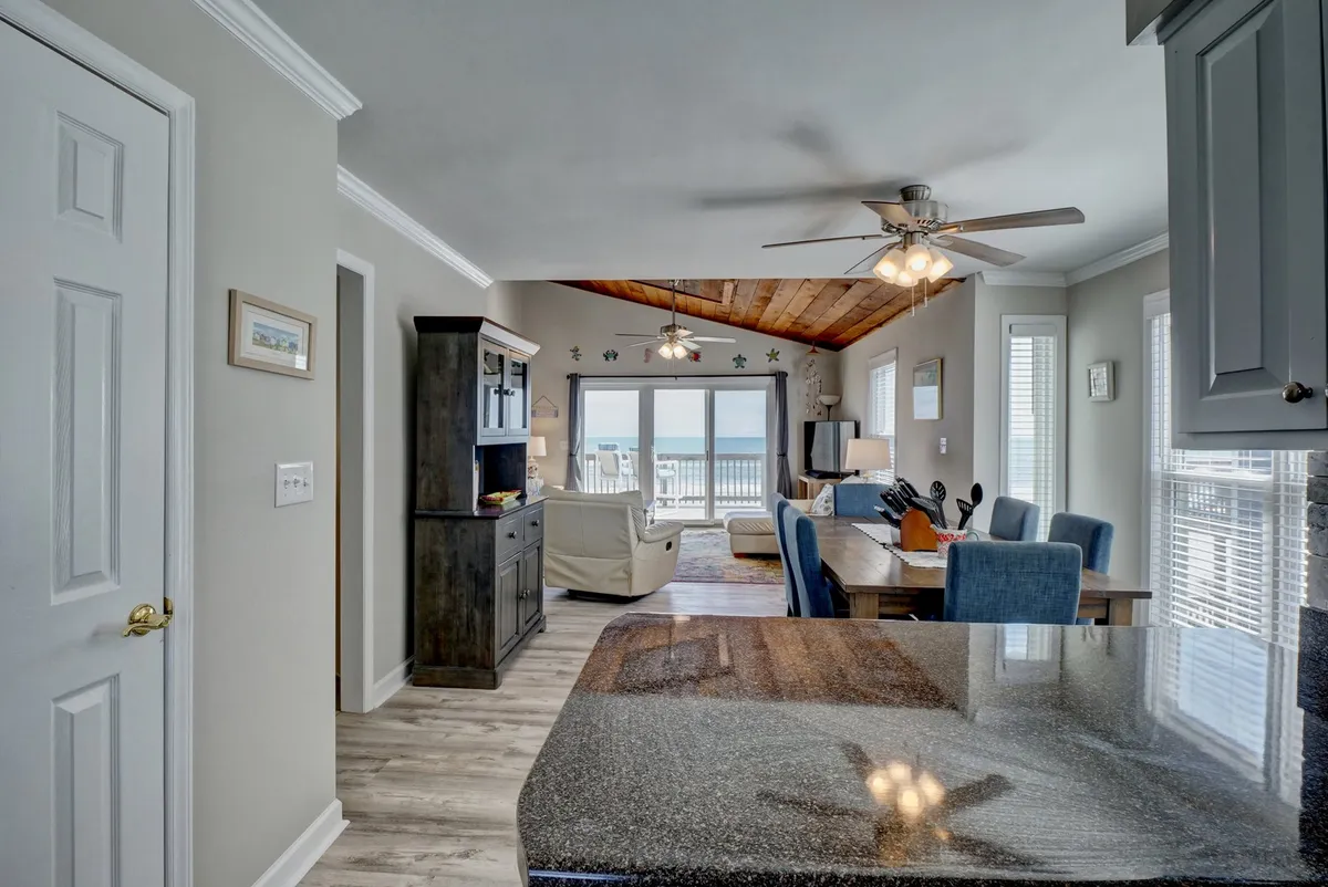This image captures a spacious, open-concept living area featuring a granite countertop that transitions seamlessly into the dining area and comfortable living room seating. Large windows offer stunning ocean views, bathing the room in natural light. The layout highlights the flow between kitchen, dining, and living space, with inviting décor that encourages gatherings.