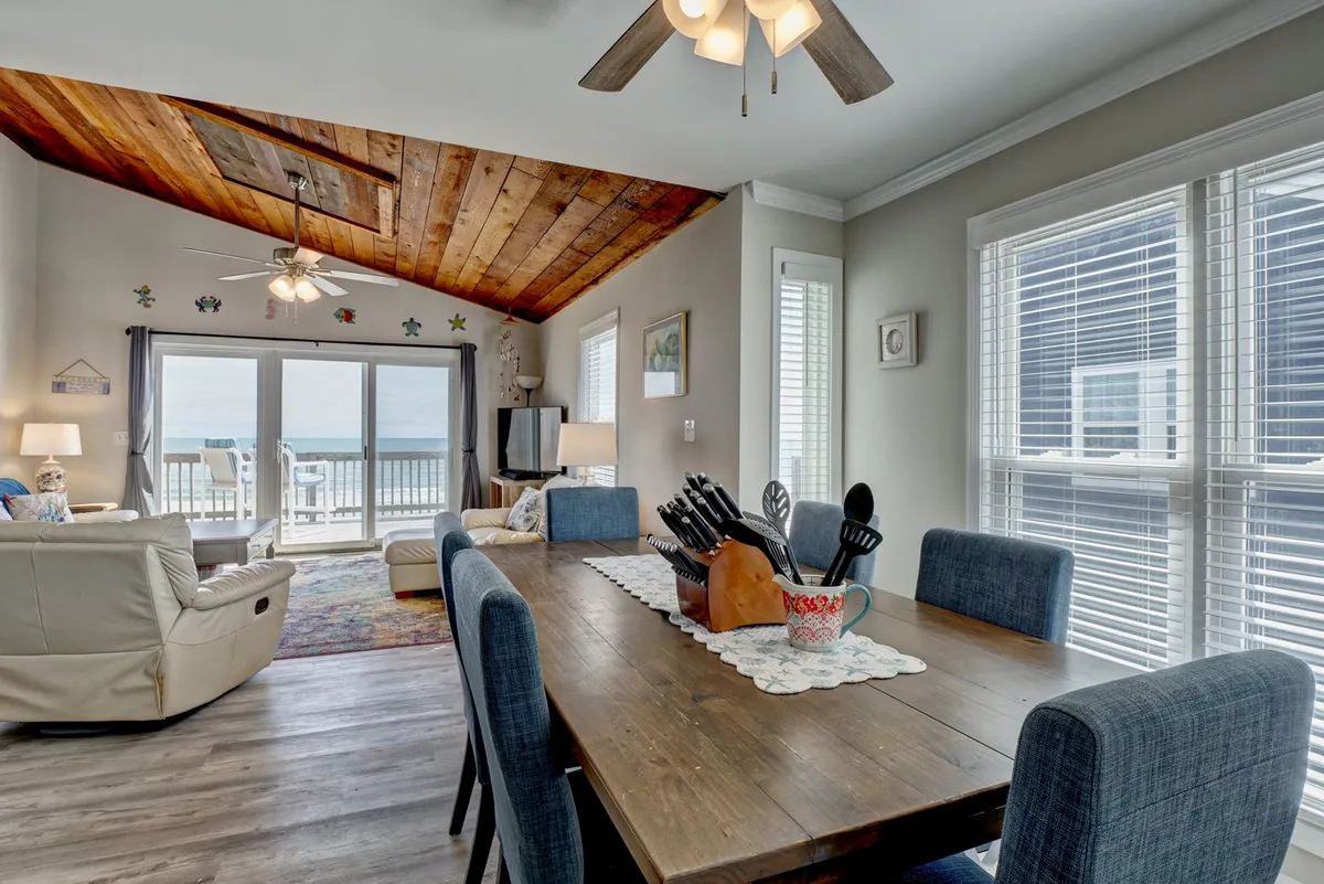 A bright and airy dining area with a wooden ceiling and large sliding glass doors. The dining table is set for meals, and the kitchen space is visible, reinforcing the theme of home-style convenience and comfort.