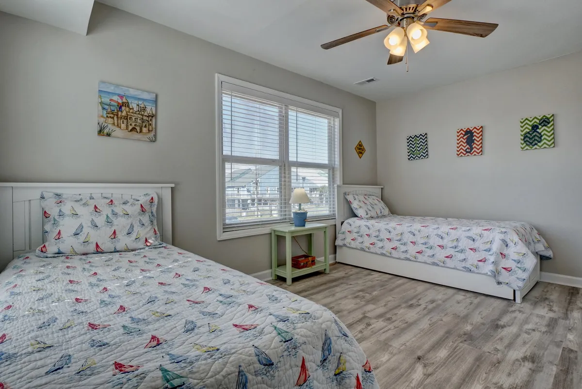 A playful, brightly lit bedroom designed for children, featuring two twin beds with nautical-themed quilts, a large window streaming in natural light, and colorful wall art. The spacious layout is ideal for kids’ activities and sleepovers, with a welcoming, safe vibe.