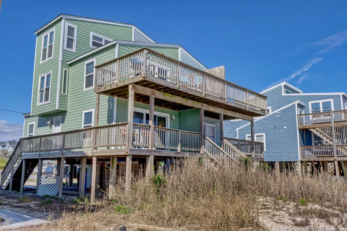 An exterior view of Sur la Mer showcasing its cheerful green siding, upper deck with seating, stilted foundation, and lush surrounding vegetation. The deck is set up for outdoor gatherings, sunbathing, or family meals, with open views ideal for relaxing and group activities.