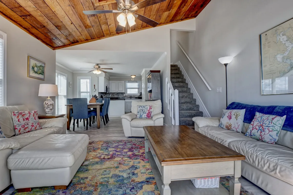 A spacious living area featuring a wooden ceiling, colorful area rug, and plush sofas arranged for socializing. The adjacent dining table is set near large windows, filling the room with natural light and offering an inviting atmosphere for gatherings.